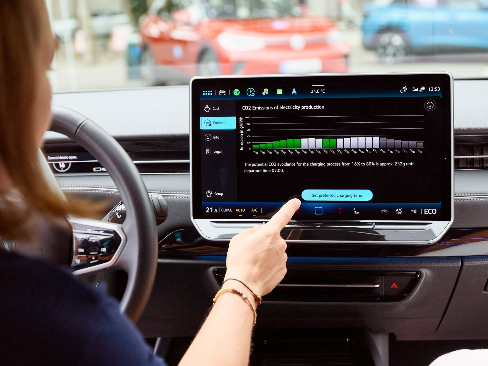 Innenansicht eines modernen Autos, in dem eine Person den großen zentralen Touchscreen bedient. Auf dem Display sind Informationen zum Ladevorgang und zur CO₂-Emission der Stromproduktion zu sehen, inklusive Diagramm und Text. Draußen vor der Windschutzscheibe stehen weitere Fahrzeuge auf einem Parkplatz oder an einer Straße.