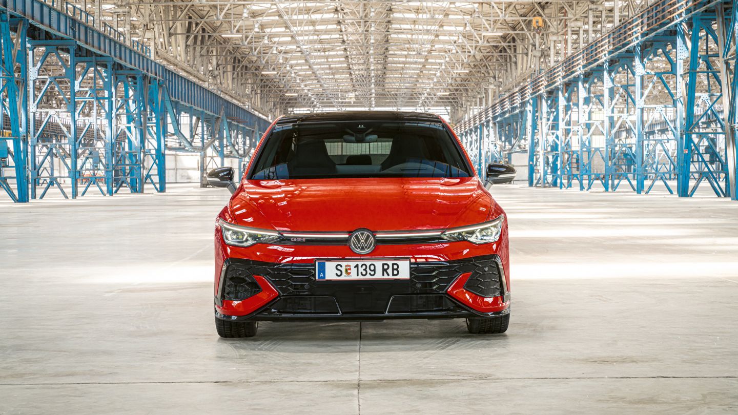 Ein roter Golf GTI steht in einer großen, hell beleuchteten Lagerhalle. Das Auto ist von vorne zu sehen und hat das Kennzeichen MÖB-GT 150. Im Hintergrund sind metallene Strukturen und Säulen zu sehen.