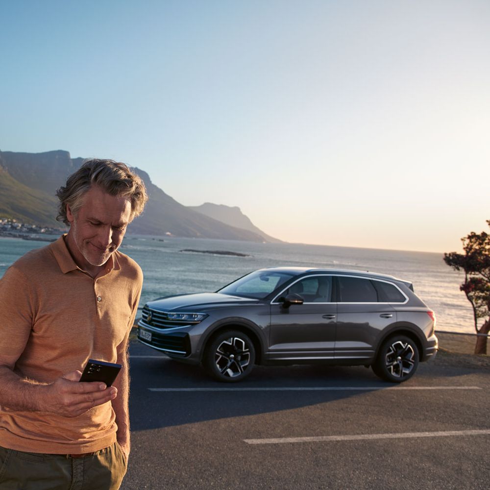 Ein Mann schaut auf sein Smartphone, dahinter parkt ein VW Touareg Elegance, im Hintergrund das Meer und die Küste.