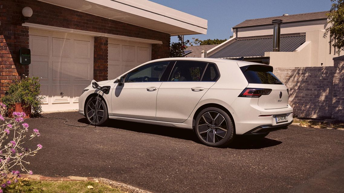 Ein weißer VW Golf eHybrid lädt an einer Ladestation vor einer Garage.