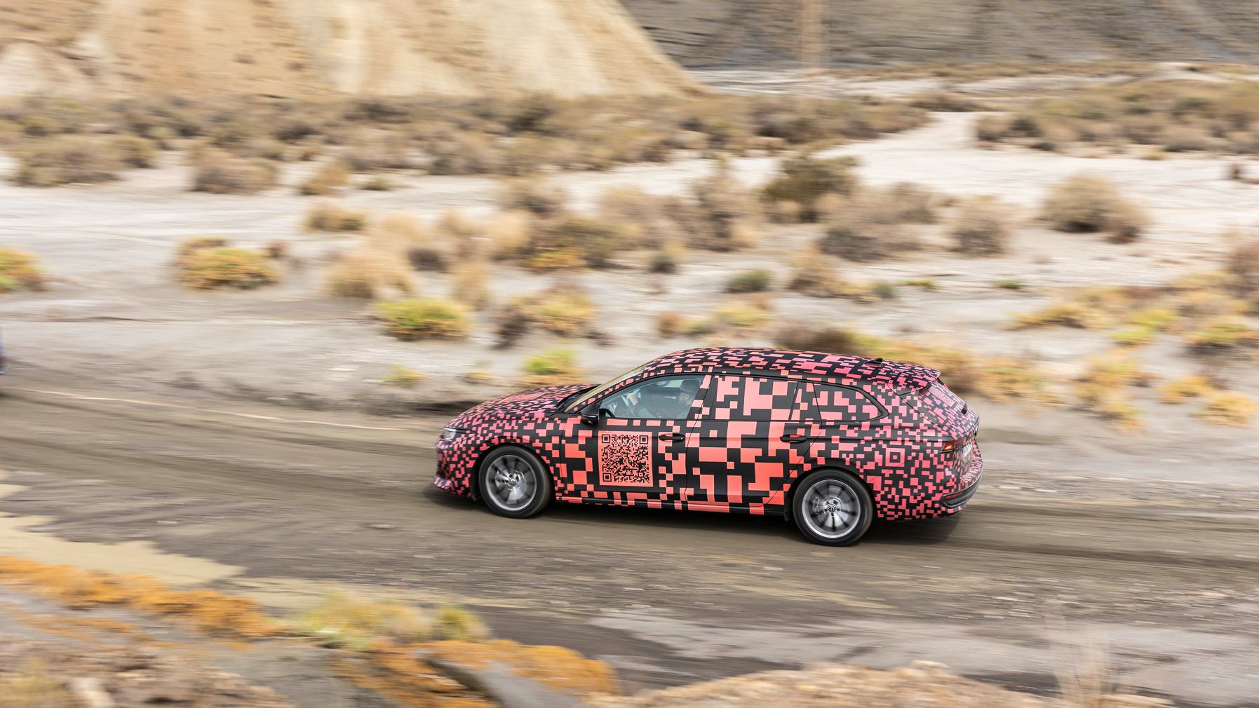 Ein Auto fährt durch eine trockene, hügelige Landschaft. Das Fahrzeug ist mit einem auffälligen, rot-schwarzen Pixelmuster beklebt. Es handelt sich vermutlich um ein Testfahrzeug.