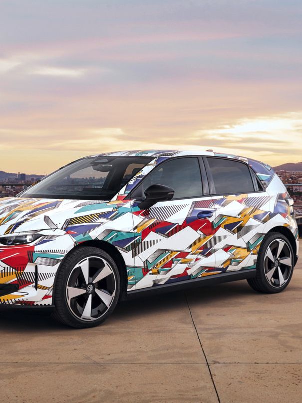 Ein bunt bemalter ID. Polo steht auf einem Parkplatz mit Stadt und Hügeln im Hintergrund. Das Fahrzeug ist mit einem lebhaften, abstrakten Design bedeckt. Der Himmel ist teils bewölkt mit Sonnenaufgang oder Sonnenuntergang Farben.