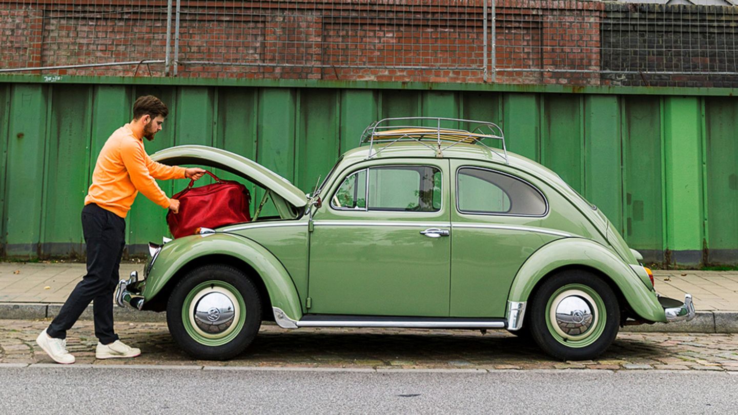 Ein Mann mit orangefarbenem Pullover lädt eine rote Reisetasche in den geöffneten Kofferraum eines hellgrünen VW Käfers an der Vorderseite. Das Auto steht am Straßenrand vor einer industriellen Kulisse mit grünen Metallwänden, Ziegelmauer und großen Rohren im Hintergrund.