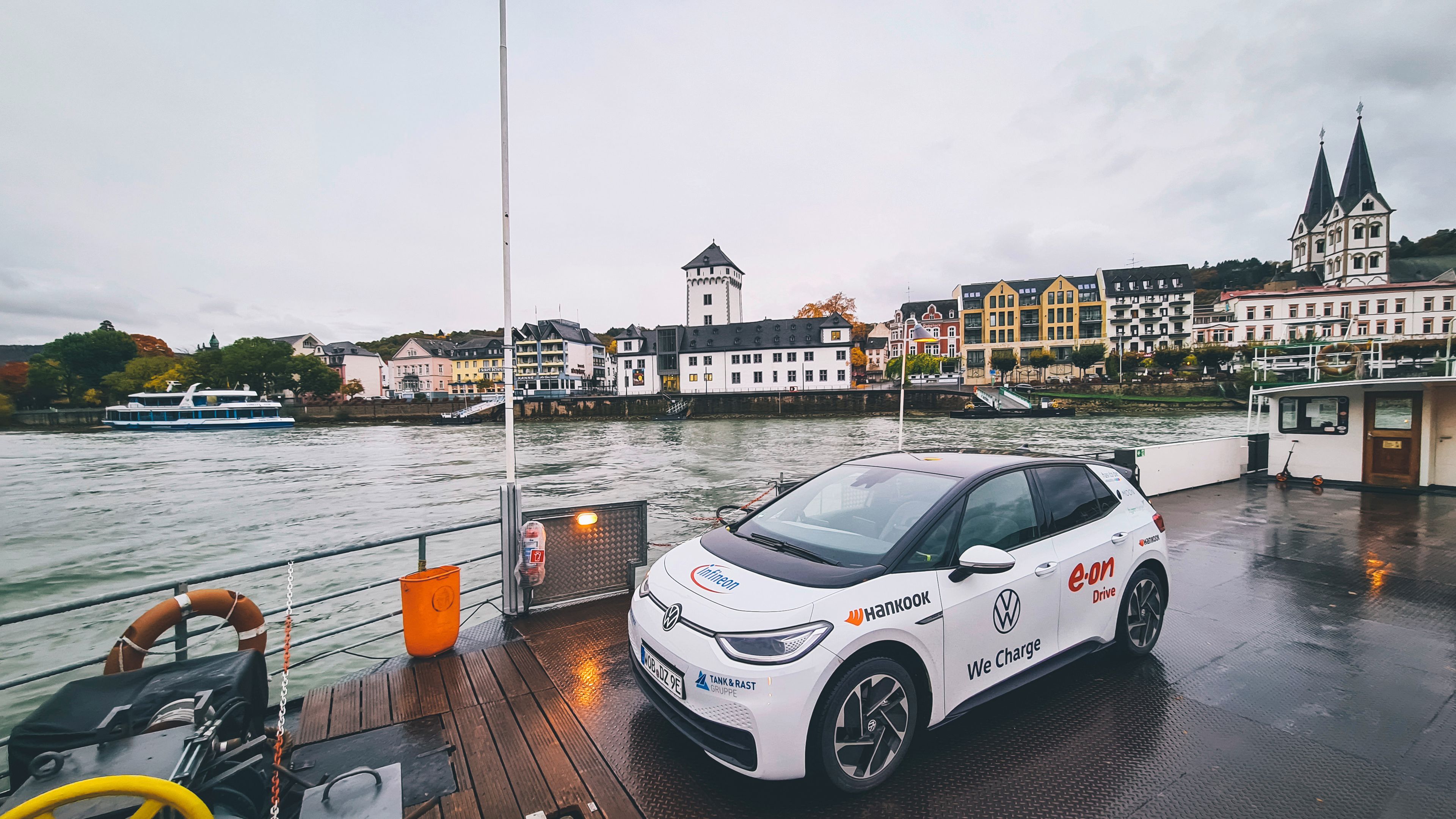 Ein weißer ID.3 steht am Deck eines Fährschiffs mit einer malerischen Stadt im Hintergrund. Das Auto ist mit Logos von Sponsoren wie VW, E.ON und Hankook bedeckt. Im Hintergrund sind ein Fluss, Häuser und eine Kirche zu sehen.