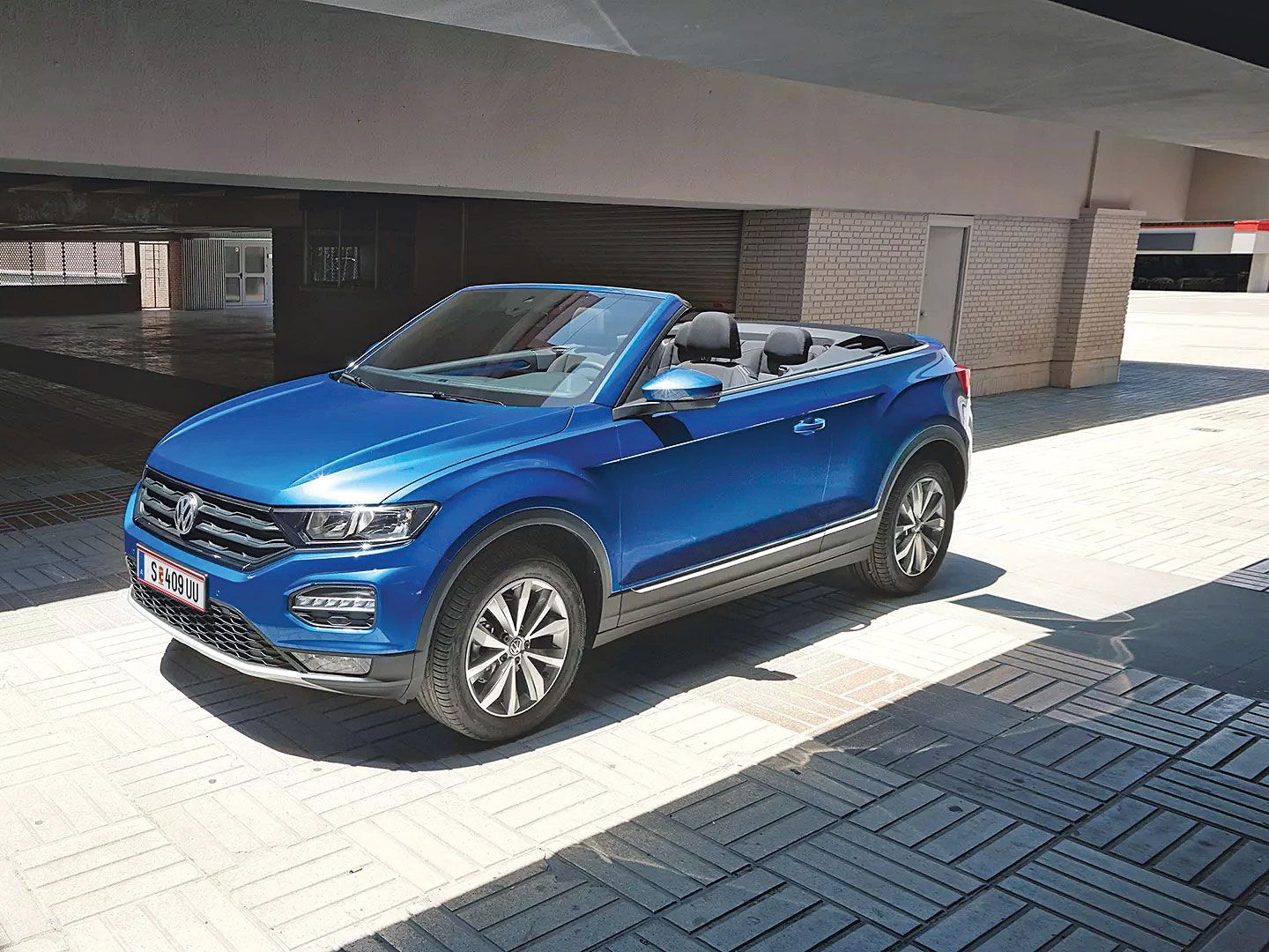 Ein blaues Volkswagen Cabriolet steht in einem sonnigen, überdachten Bereich. Es hat das Verdeck geöffnet. Die Umgebung ist modern und urban.