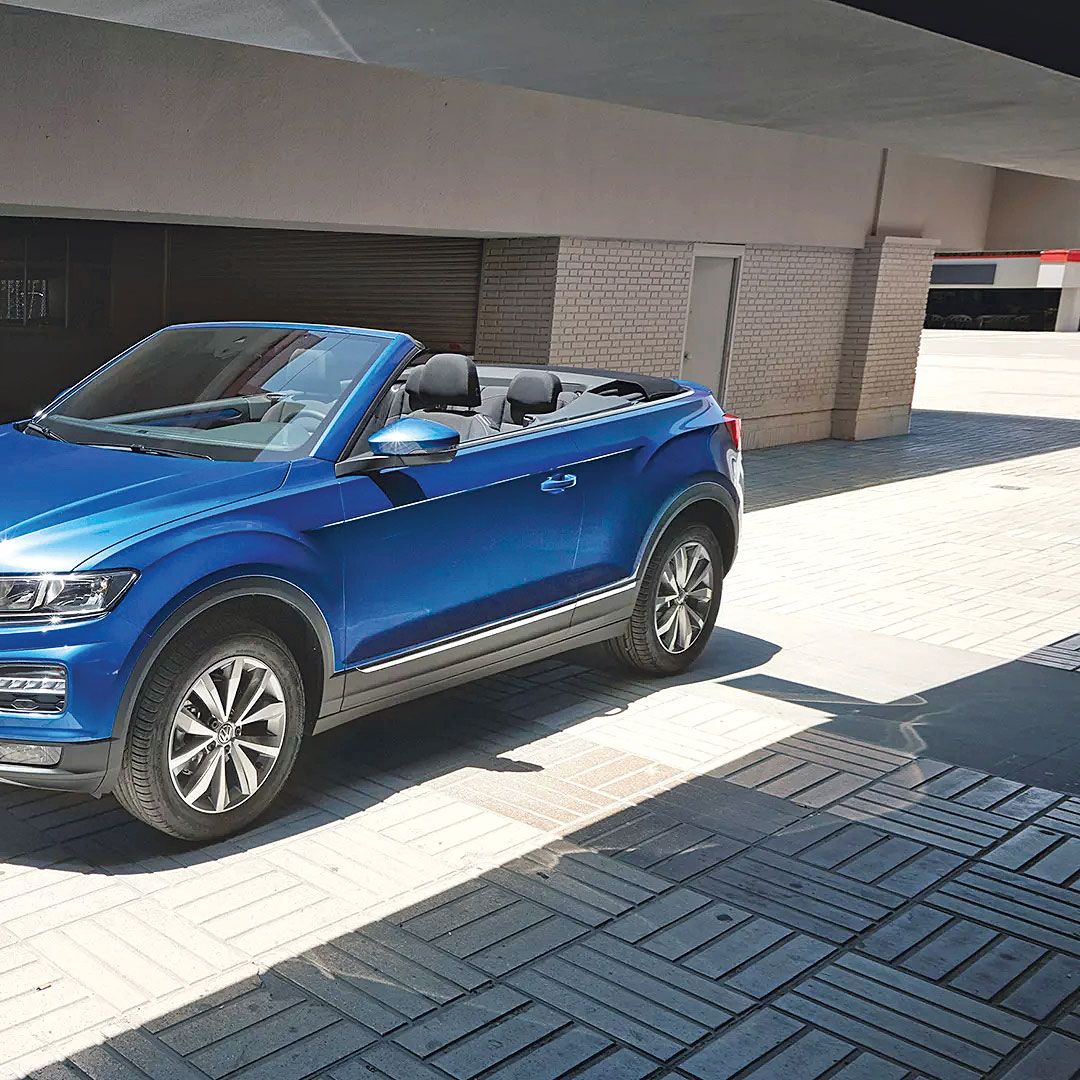 Ein blaues Volkswagen Cabriolet steht in einem sonnigen, überdachten Bereich. Es hat das Verdeck geöffnet. Die Umgebung ist modern und urban.