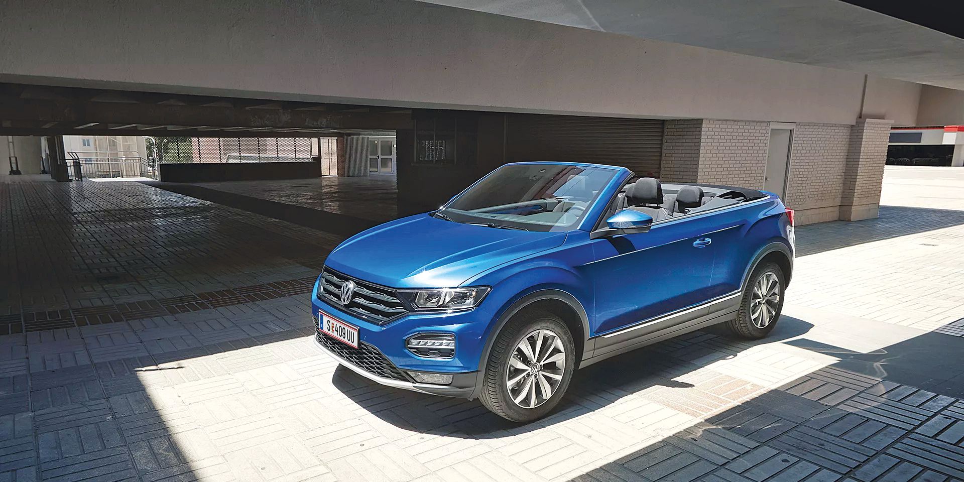 Ein blaues Volkswagen Cabriolet steht in einem sonnigen, überdachten Bereich. Es hat das Verdeck geöffnet. Die Umgebung ist modern und urban.