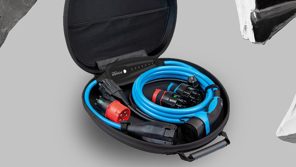 Ein offener, ovaler Koffer enthält blaue und schwarze Kabel sowie Anschlüsse. Der Hintergrund ist grau und mit schwarz-weißen Gesteinsformen verziert. Das Bild zeigt Ausrüstung für den Anschluß von Elektrofahrzeugen.