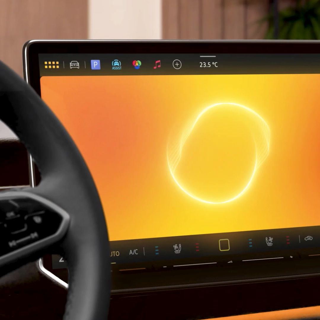 Ein modernes Auto-Armaturenbrett mit einem Bildschirm, der eine hellgelbe Oberfläche und verschiedene Icons am oberen Rand anzeigt. Links ist teilweise das Lenkrad zu sehen. Das Display zeigt auch die Temperatur und Klimaanlagen-Einstellungen an.