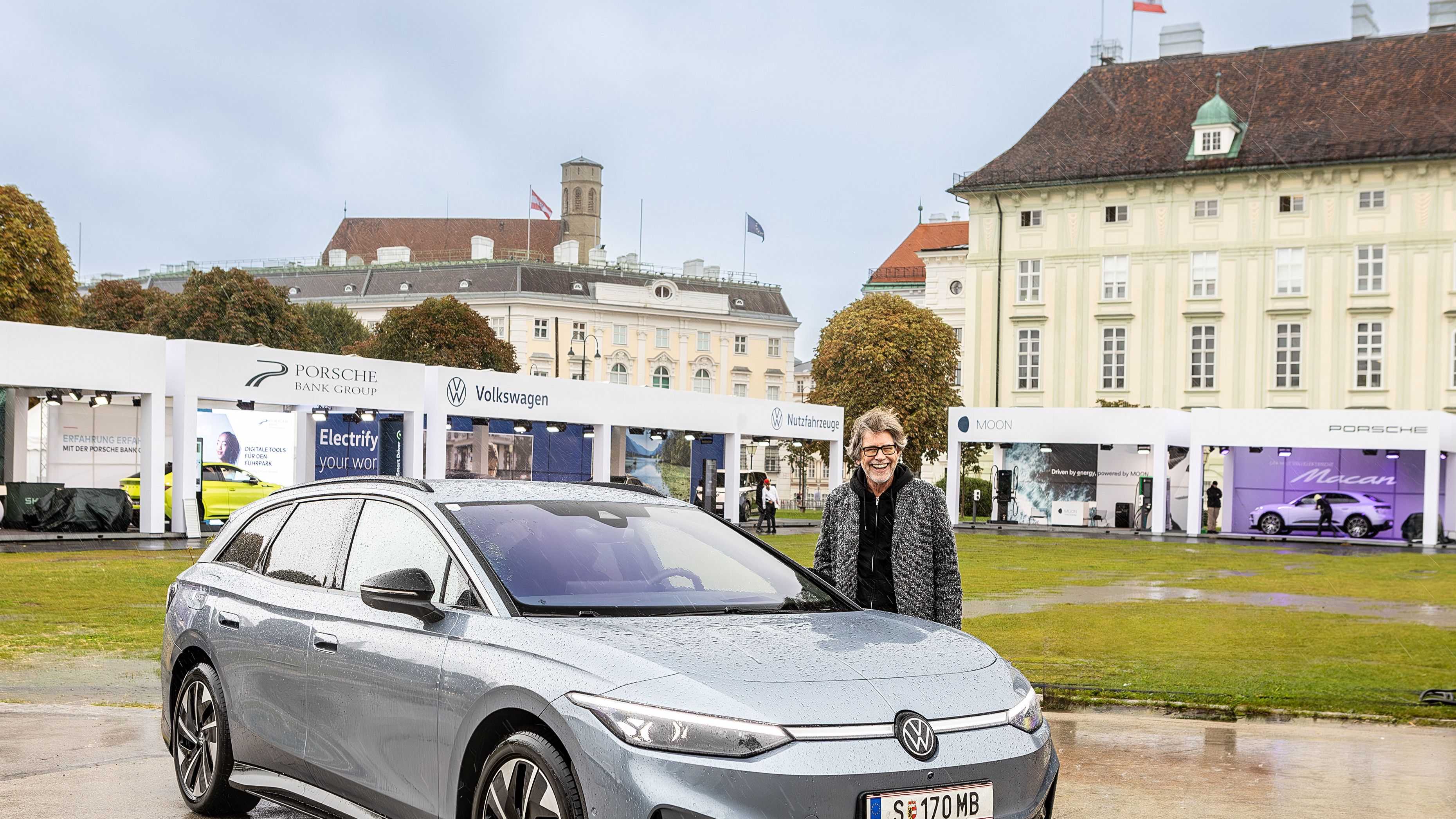 Ein silberner Volkswagen steht vor einem historischen Gebäude auf einem nassen Platz. Im Hintergrund sind Stände von Porsche und Volkswagen zu sehen. Ein Mann steht neben dem Auto.