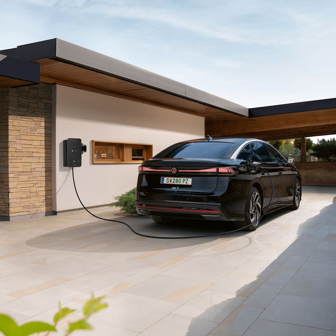 Ein schwarzes Elektroauto steht in einer modernen Einfahrt und ist an einer Wallbox an der Hauswand zum Laden angeschlossen. Das flache, kubische Haus mit Holz- und Steinelementen wirkt modern und hochwertig. Im Vordergrund sind Pflanzen zu sehen, im Hintergrund öffnet sich der Blick auf eine Landschaft.