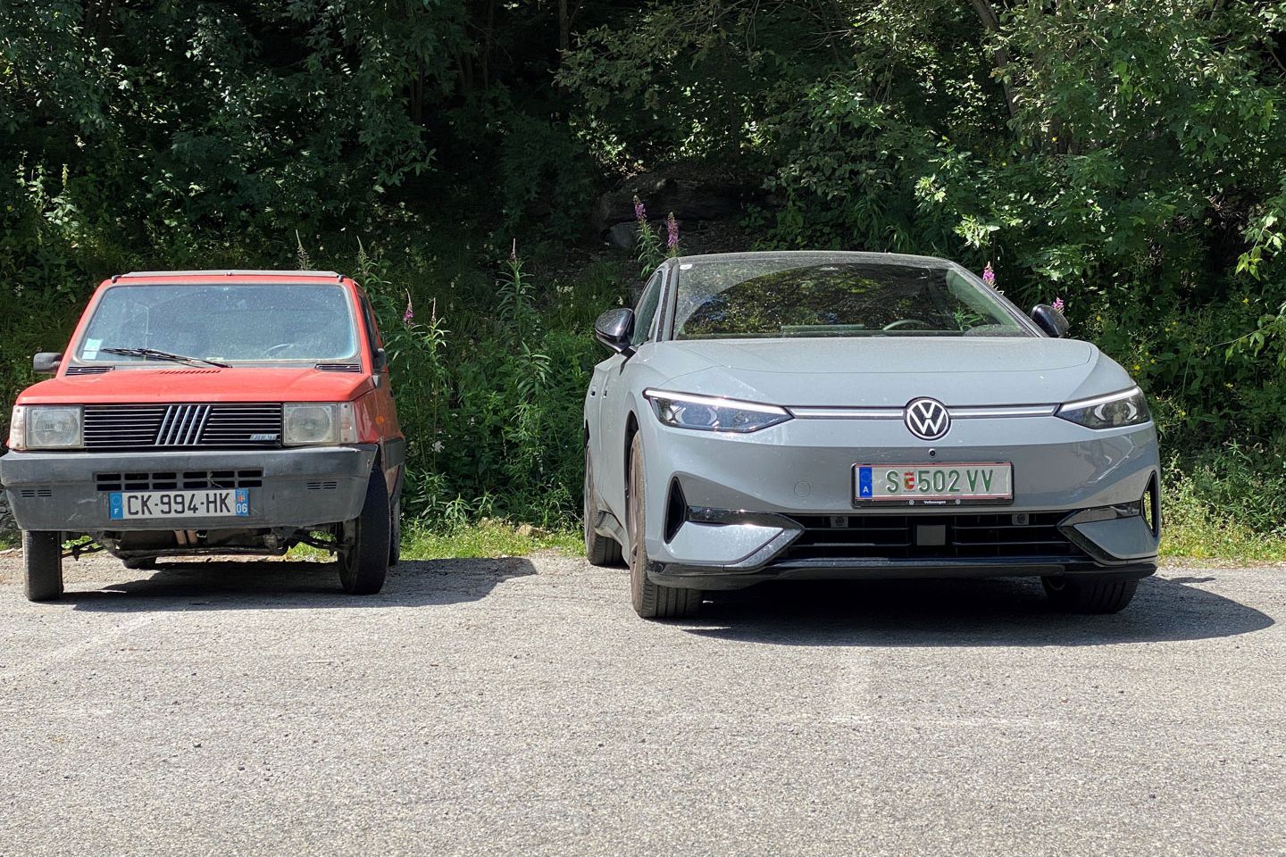 Ein rotes Fiat-Auto und ein graues Volkswagen-Elektroauto stehen nebeneinander auf einem Parkplatz. Das Fiat-Auto ist älter und kleiner, während das Volkswagen-Auto größer und moderner ist. Im Hintergrund sind Bäume und Vegetation zu sehen.