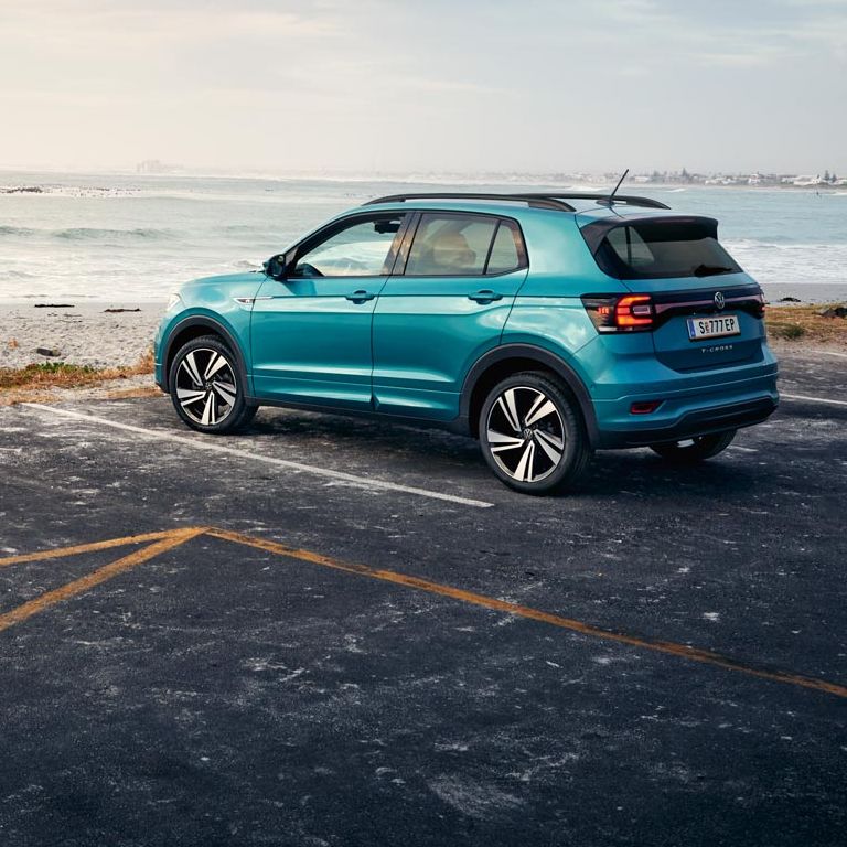  VW T-Cross R-Line in Makena-Türkis Metallic auf asphaltiertem Parkplatz an Küste mit kieseligem Strand, Blick von hinten links