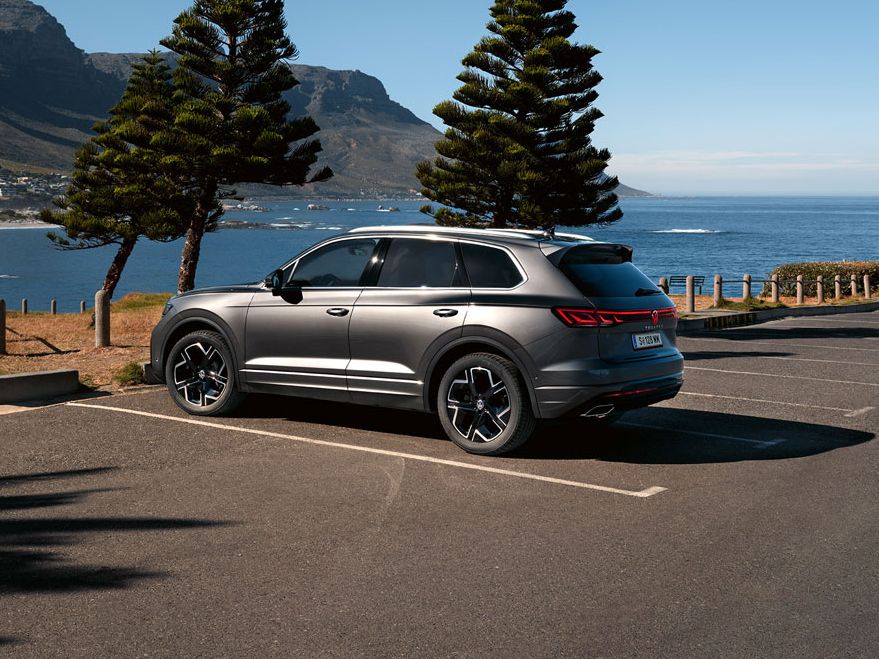 Ein VW Touareg Elegance parkt in der Sonne, Ansicht von der Seite, dahinter sind das Meer und die Küste zu sehen.