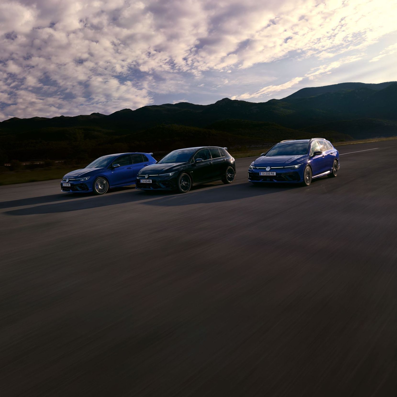 Drei VW Golf R fahren nebeneinander auf einer Straße bei Sonnenuntergang. Die Himmel zeigt einige Wolken und im Hintergrund sind Berge sichtbar. Die Farben der Autos sind blau, schwarz und dunkelblau.
