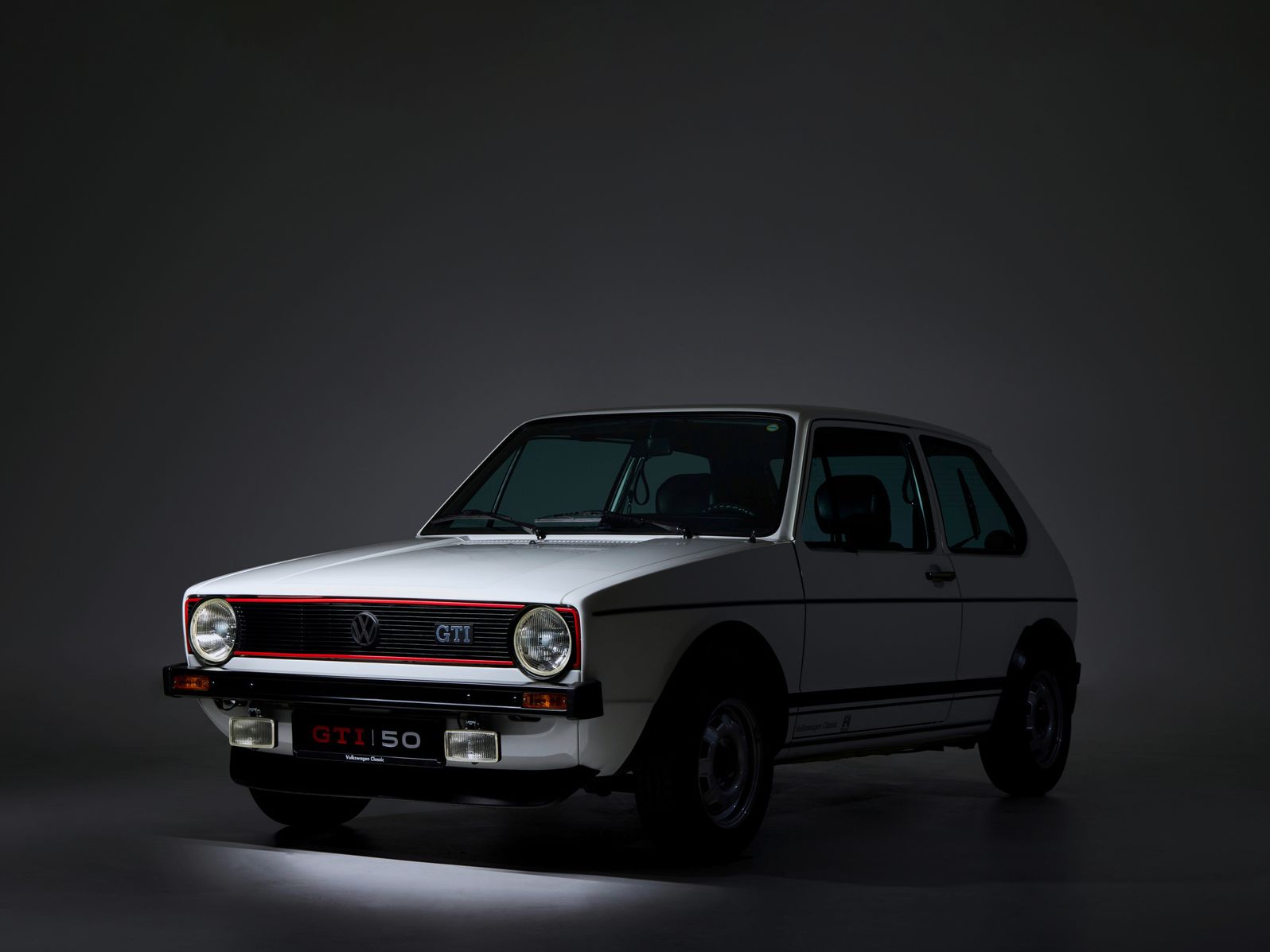 Ein weißer VW Golf GTI der ersten Generation steht in einem dunklen Studio. Das Auto ist seitlich zur Kamera ausgerichtet und hat rote und schwarze Akzente an der Front. Im Hintergrund ist nur eine schwache Beleuchtung sichtbar.