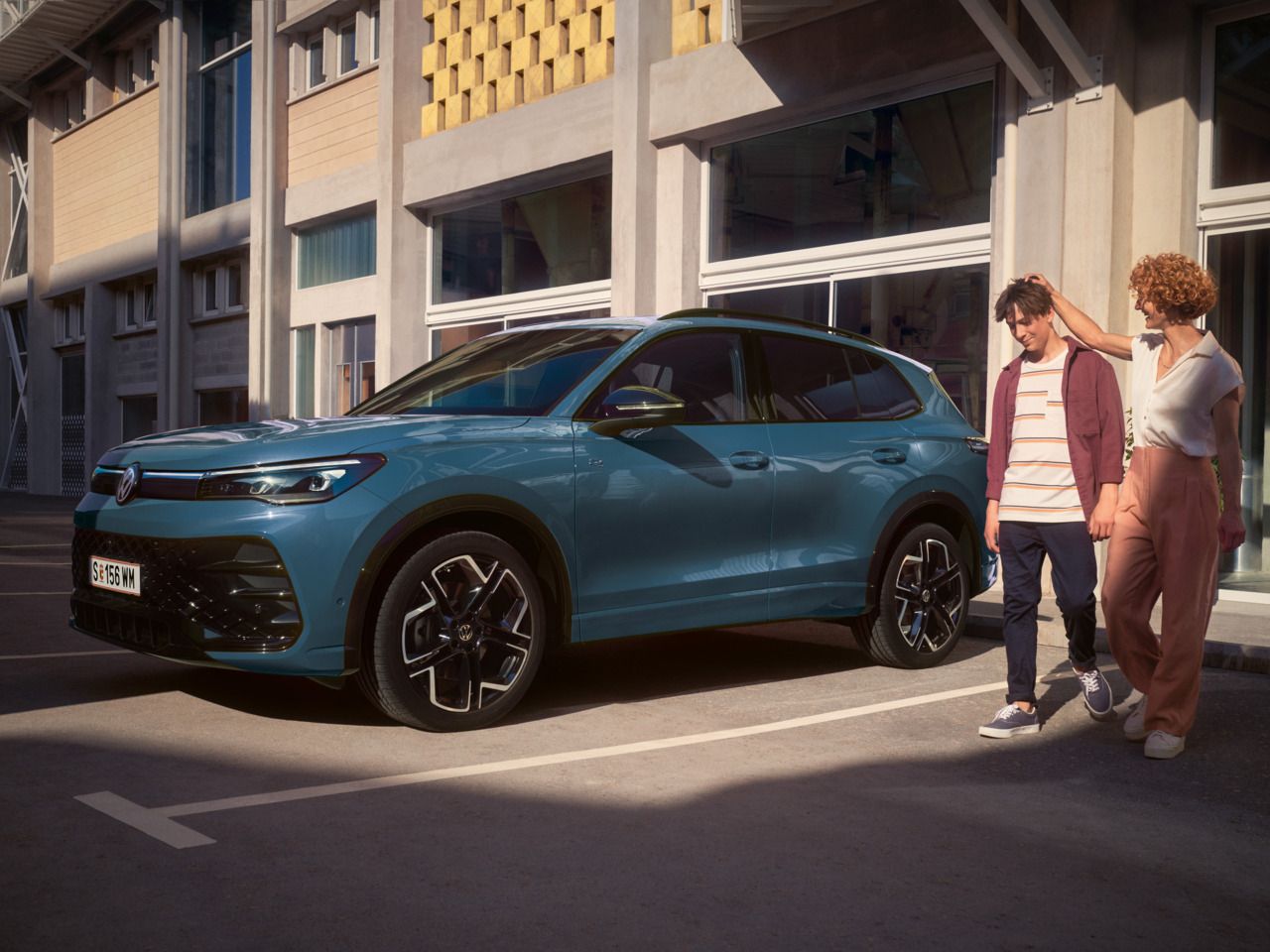 Eine Frau und ein Mann verlassen ein Gebäude mit großen Glasfenstern und gehen an einem roten VW Tiguan vorbei.