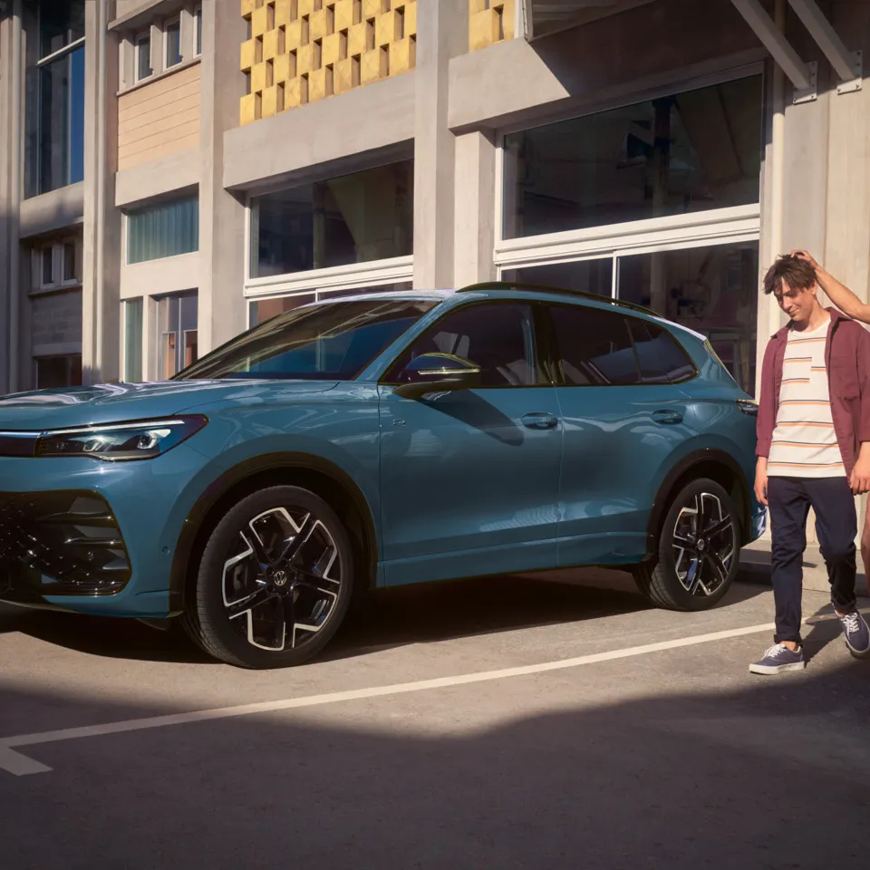 Eine Frau und ein Mann verlassen ein Gebäude mit großen Glasfenstern und gehen an einem roten VW Tiguan vorbei.