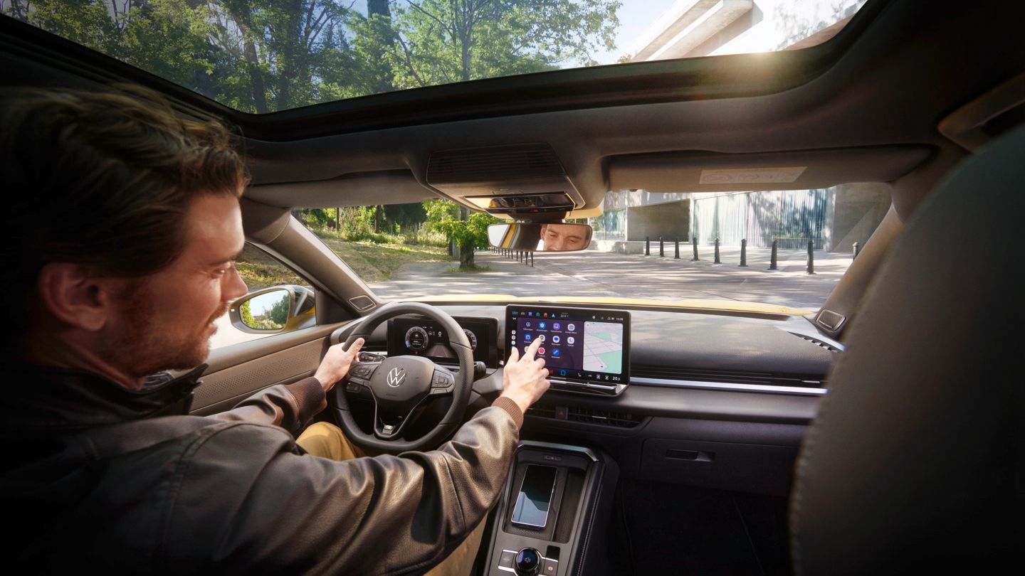 Ein Mann mit braunen Haaren und schwarzer Jacke sitzt am Fahrersitz im Interieur eines VW T-Rocs und bedient das Infotainmentsystem und mit der anderen Hand das Lenkrad. Er befindet sich auf einer Straße, im Hintergrund sieht man rechts ein modern wirkendes Gebäude, links ein bewaldetes Stück.