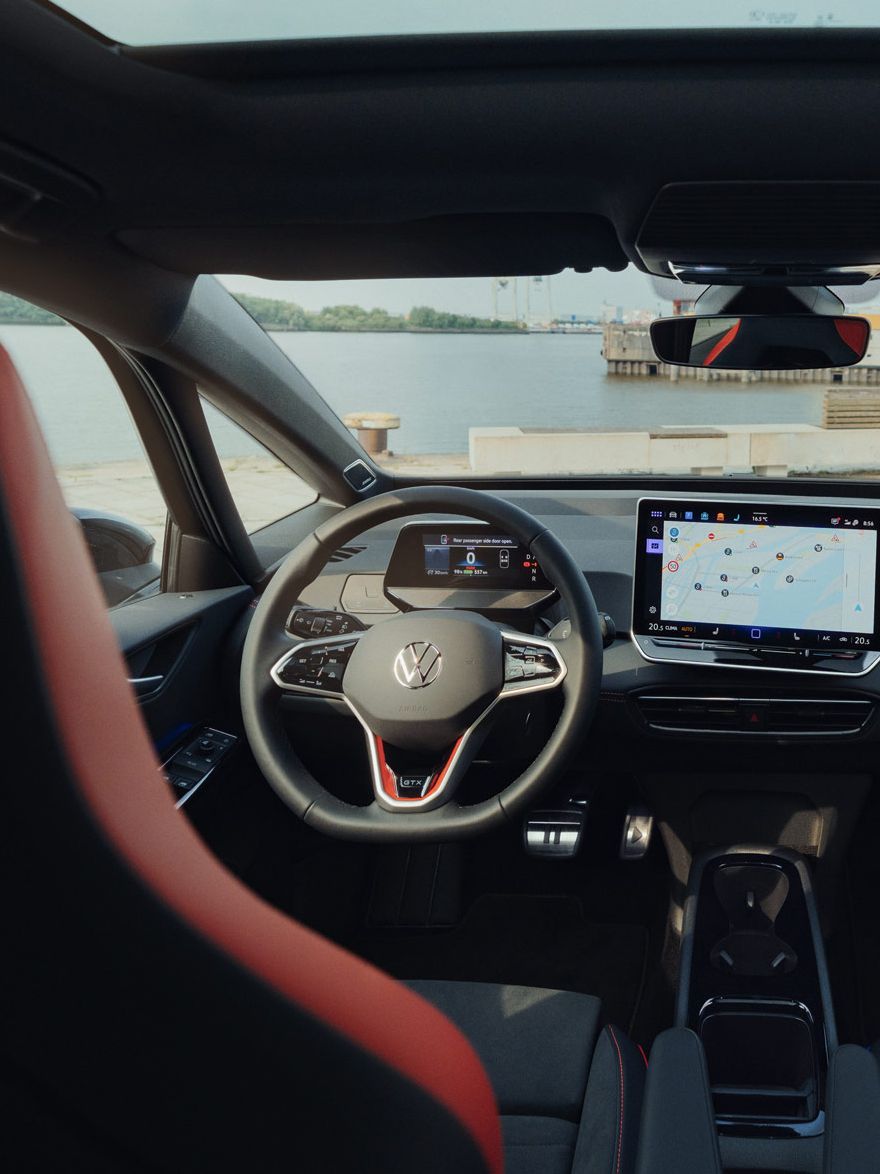 Innenraum des ID.3 GTX FIRE+ICE, das mit einem großen Touchscreen-Navigationsgerät ausgestattet ist. Zwei vordere Sitze, einer in Rot und einer in Blau, sind sichtbar. Die Aussicht aus dem Fenster zeigt eine Uferlandschaft.