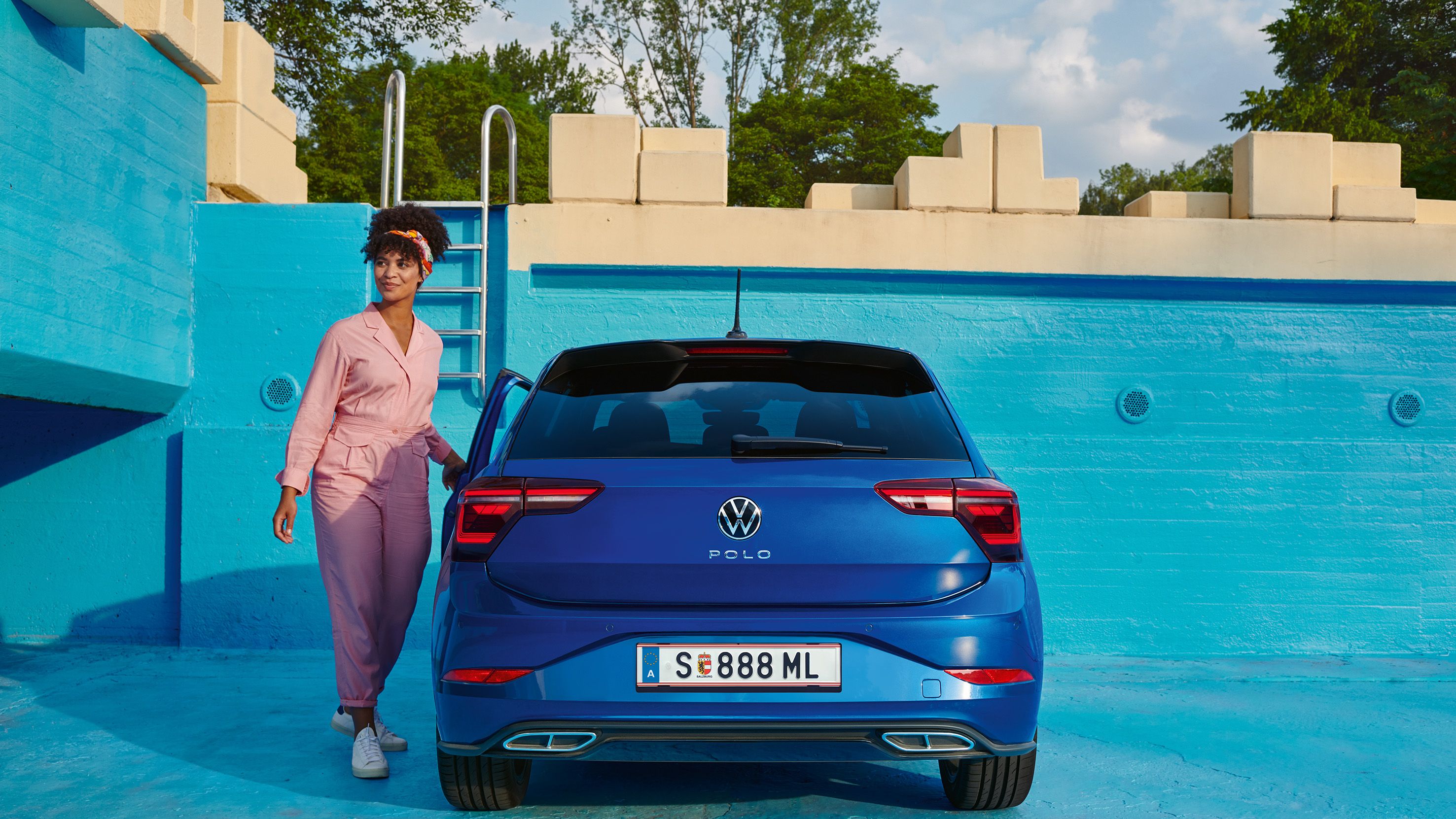 Ein blauer VW Polo steht in einem blau angemalten, leeren Poolbecken, eine junge Frau steigt ein.