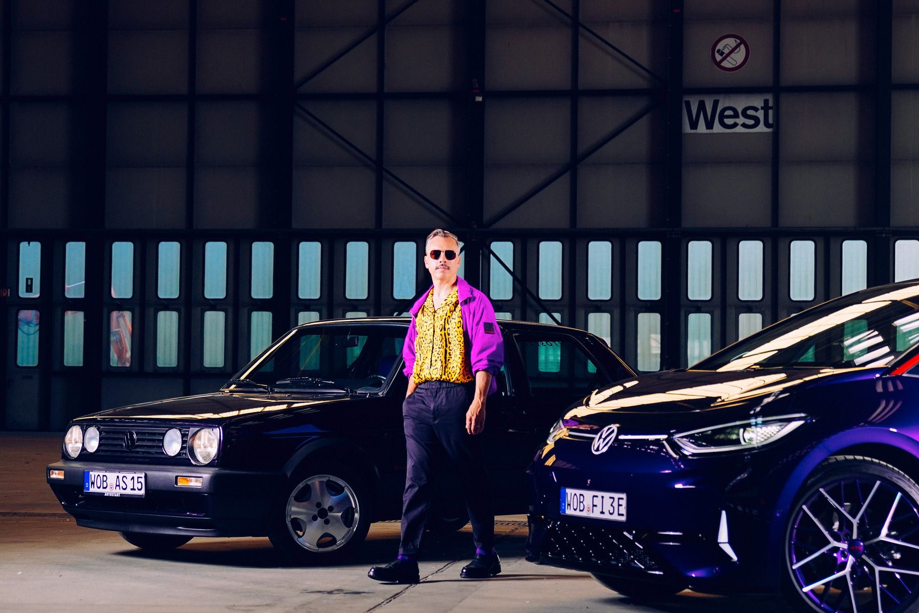 Ein Mann steht in einer Halle zwischen zwei Autos, einem schwarzen älteren Modell und einem modernen blauen Modell. Das Nummernschild der Autos zeigt abgekürzte Städte, vermutlich deutsche Städte. Die Halle hat große Fenster und Türen, die teilweise beleuchtet sind.