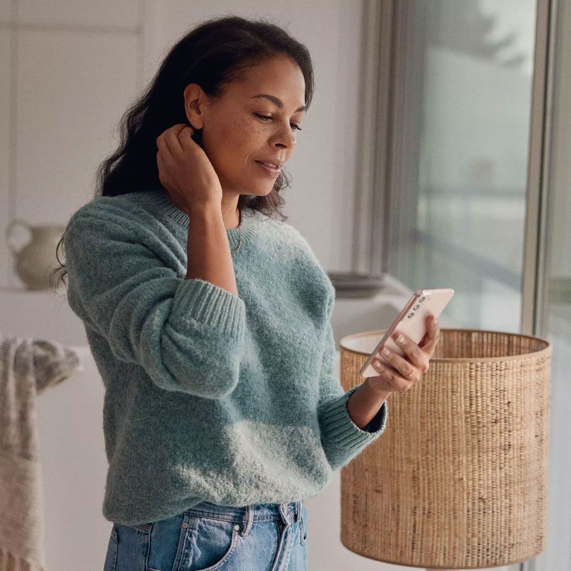 Frau in einem hellblauen Pullover und Jeans hält ein Smartphone in der Hand. Sie steht in einem Raum mit einem Fenster im Hintergrund und einem Korb auf einem Tisch. Ihr Gesicht ist unscharf/blurry.