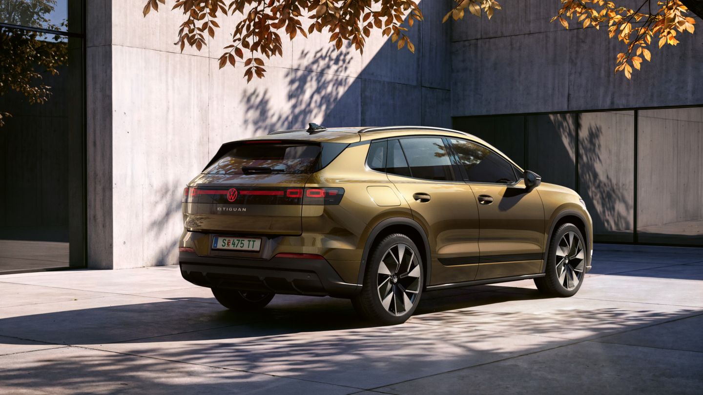 Seitliche Heckansicht eines oliv-bronzefarbenen VW ID. Tiguan geparkt vor einem modernen Wohngebäude. Ein Mann geht mit einer Fotokamera in der Hand auf das Fahrzeug zu.