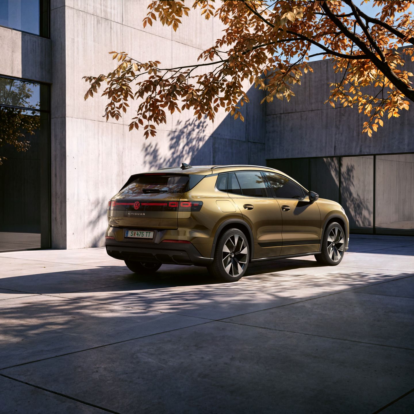 Seitliche Heckansicht eines oliv-bronzefarbenen VW ID. Tiguan geparkt vor einem modernen Wohngebäude. Ein Mann geht mit einer Fotokamera in der Hand auf das Fahrzeug zu.