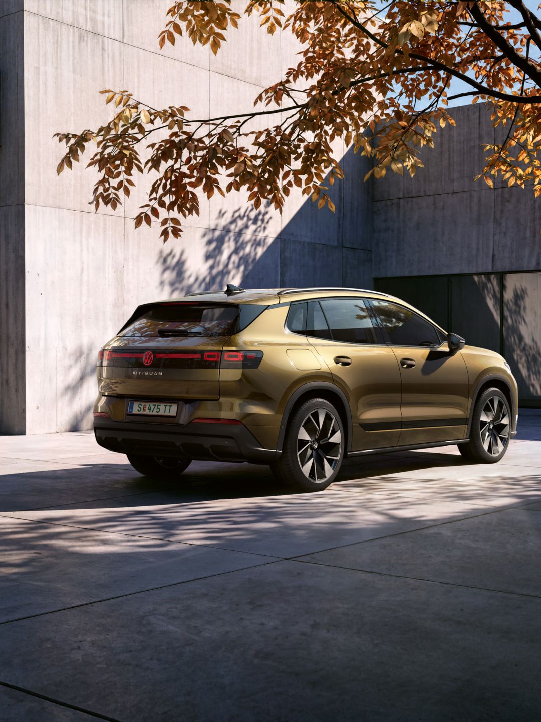 Seitliche Heckansicht eines oliv-bronzefarbenen VW ID. Tiguan geparkt vor einem modernen Wohngebäude. Ein Mann geht mit einer Fotokamera in der Hand auf das Fahrzeug zu.