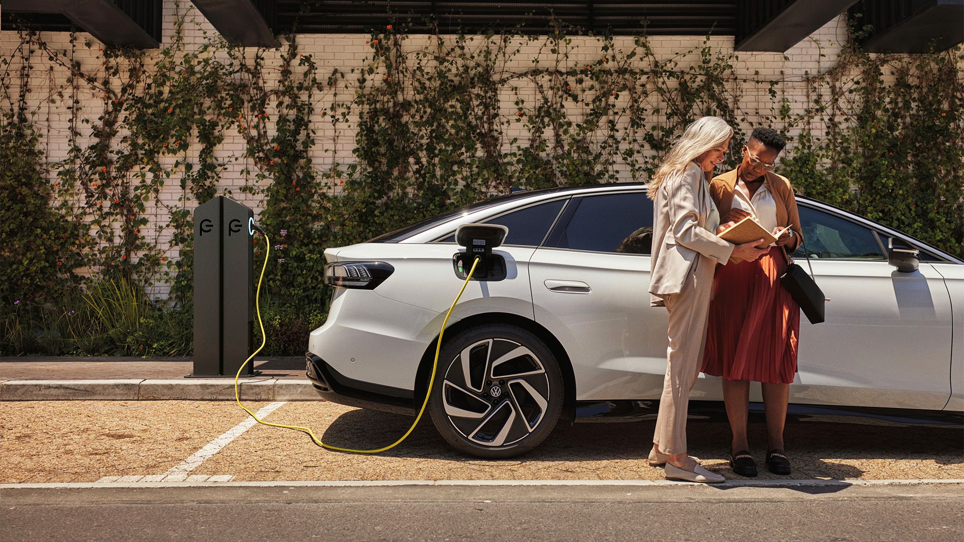 Zwei Frauen stehen mit einem Buch in der Hand neben einem weißen VW ID., der an einer öffentlichen Ladesäule lädt.