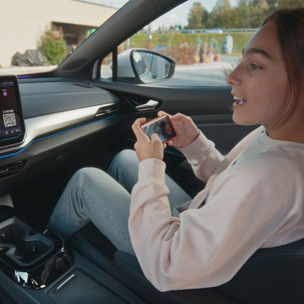 Eine Person sitzt entspannt im Fahrersitz eines Autos und spielt ein Videospiel auf ihrem Smartphone. Das Auto hat ein großes eingebautes Display auf dem Armaturenbrett, das ebenfalls ein Videospiel zeigt. Im Hintergrund ist die Außenseite eines Gebäudes zu sehen.