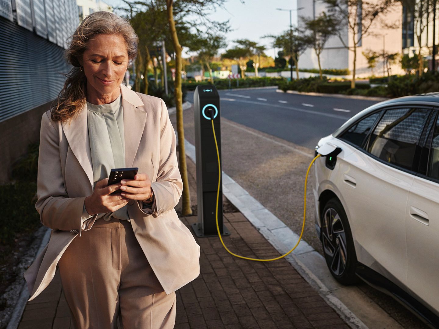 Weißer VW ID.7 seitlich lädt an einer öffentlichen Ladesäule. Eine Frau geht mit dem Smartphone in der Hand daran vorbei.