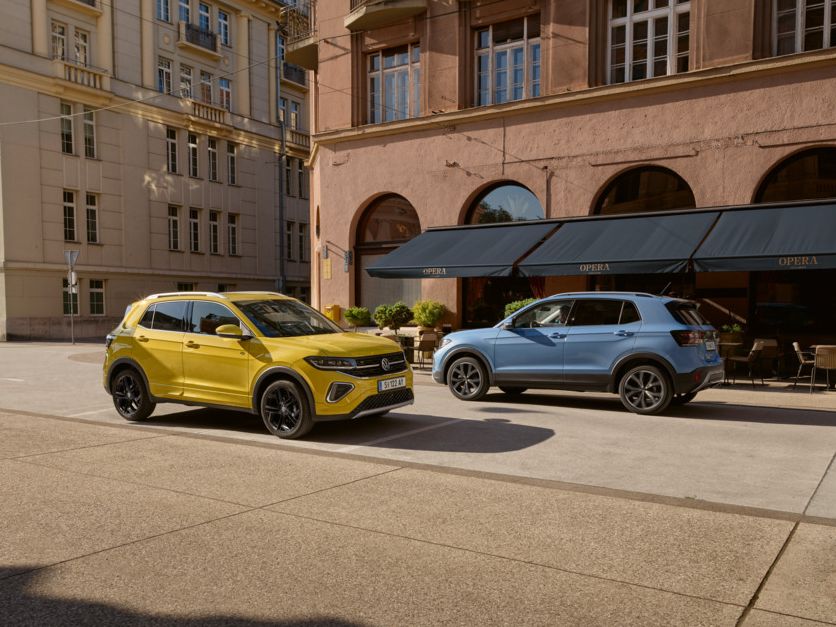 Ein gelber VW T-Cross parkt am Straßenrand in einer Stadt. Ein blauer T-Cross fährt daran vorbei.
