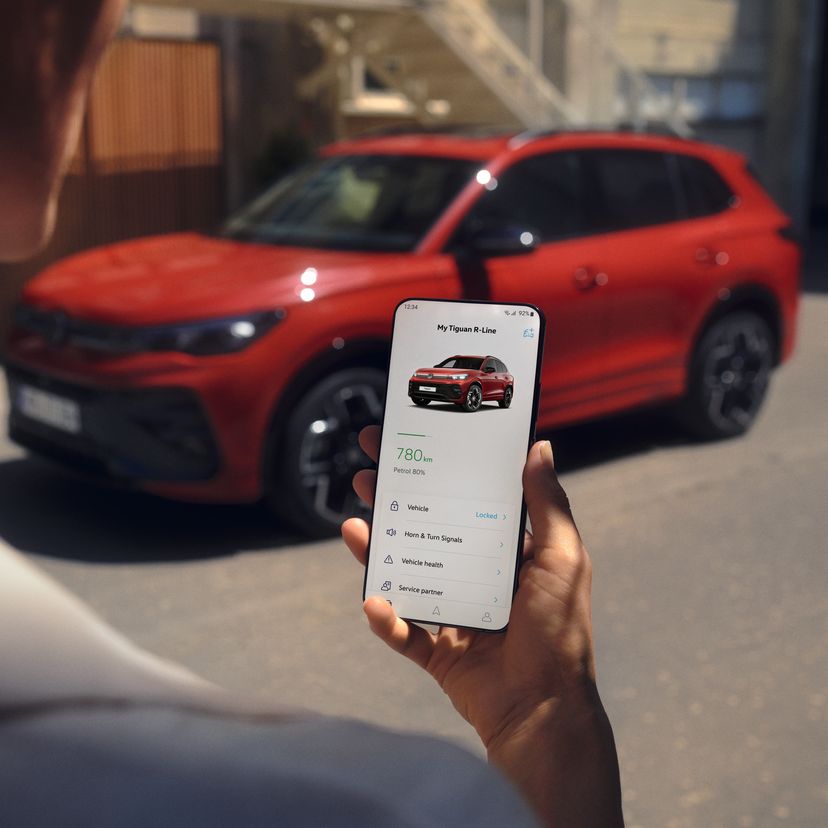 Blick über die Schulter einer Person auf ein Handy, auf dem die Volkswagen App geöffnet ist. Im Hintergrund steht ein roter Tiguan R-Line.