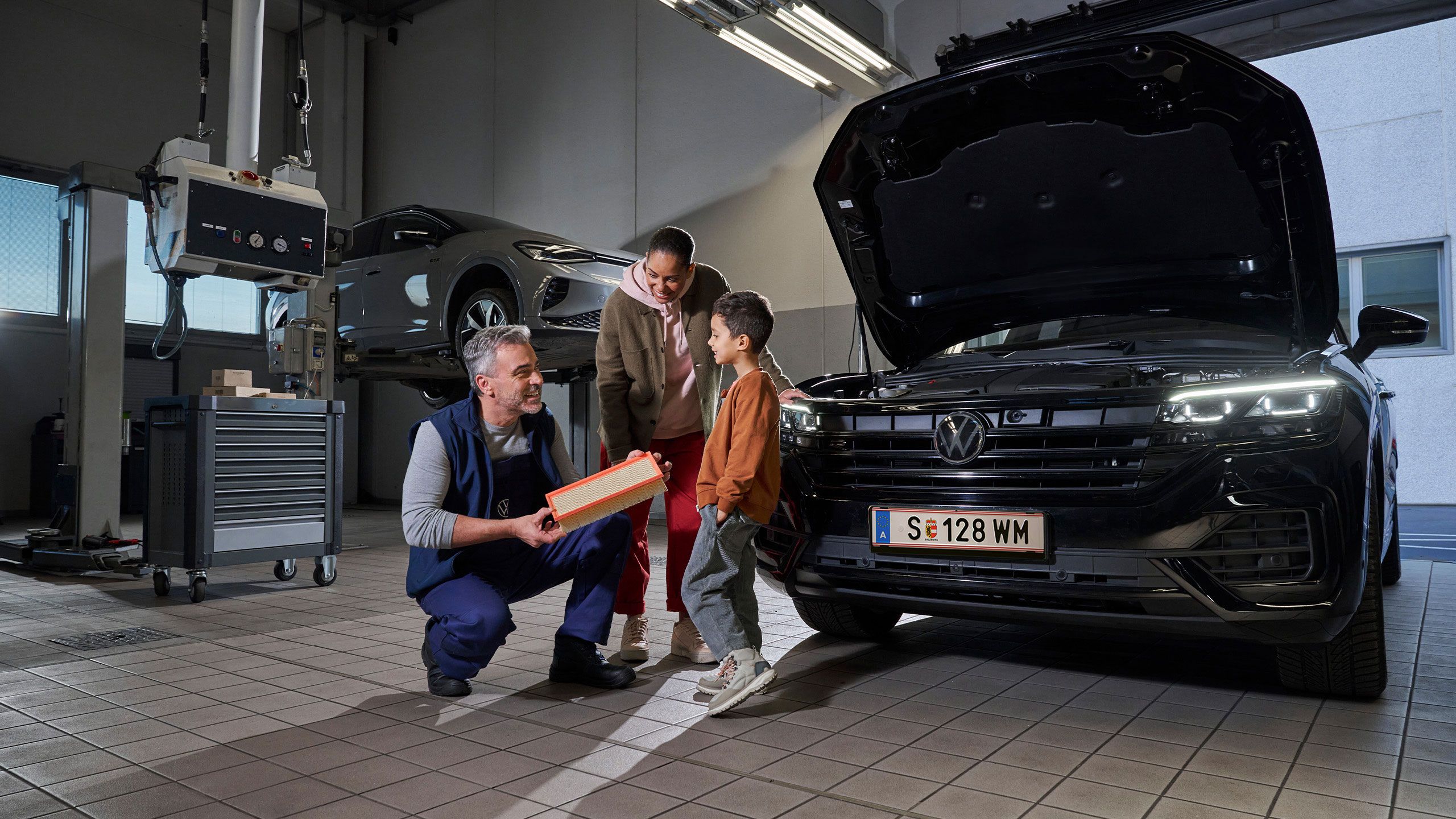 Ein Mechaniker erklärt den Aufbau eines Filters in einer Autowerkstatt. Im Hintergrund sind ein angehobenes Auto und ein geöffneter Motorraum eines schwarzen Touareg