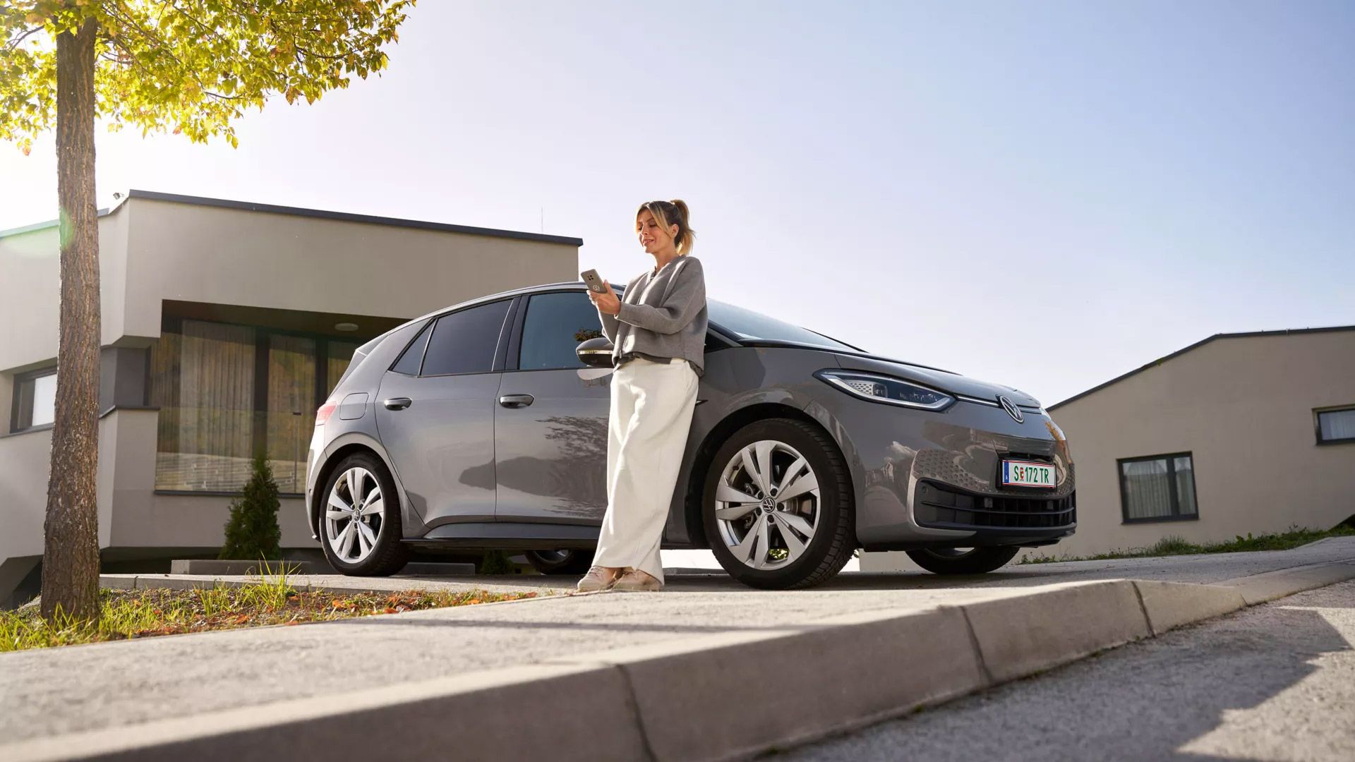 Eine Person steht neben einem grauen Elektroauto auf einer gepflasterten Auffahrt vor modernen Häusern. Sie scheint ein mobiles Gerät zu benutzen. Im Hintergrund sind Bäume und eine klare Himmel sichtbar.