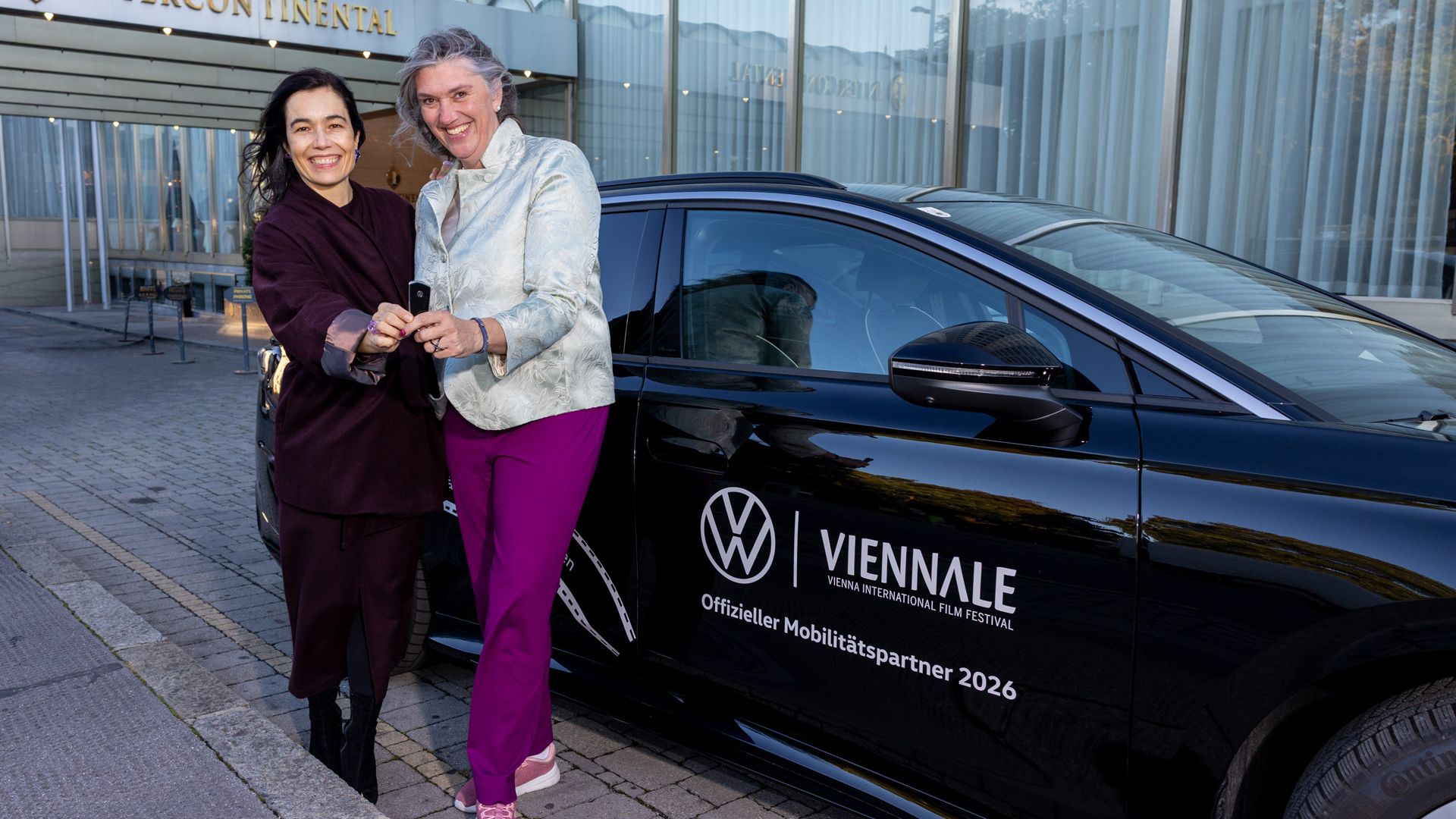 Zwei lächelnde Frauen stehen vor einem schwarzen Auto mit der Aufschrift „Viennale – offizieller Mobilitätspartner 2026“ und tauschen einen Autoschlüssel. Im Hintergrund ist der Eingang eines Intercity-Hotels mit Glasfassade zu sehen.