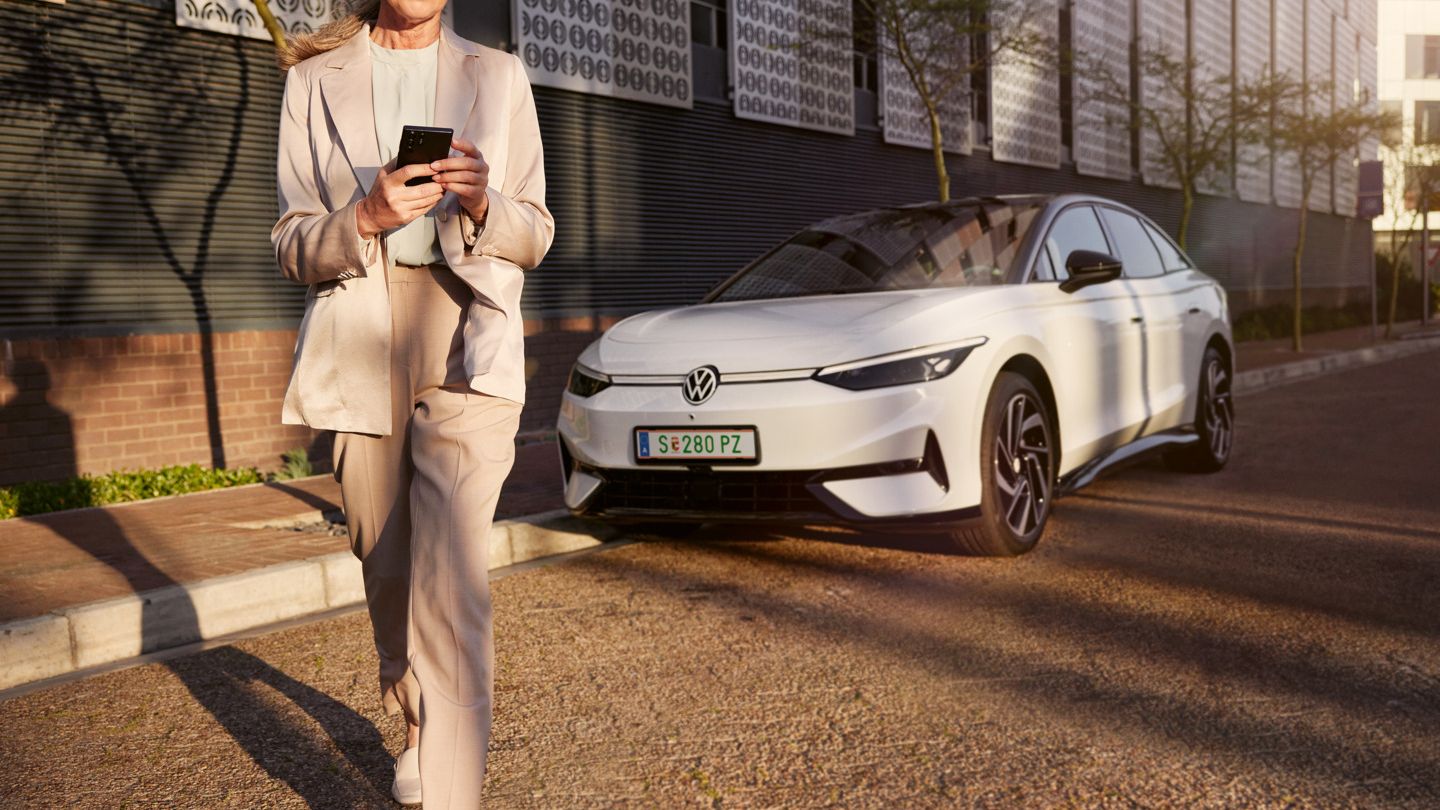 Eine Frau im Anzug schaut auf ihr Smartphone, im Hintergrund parkt ein weißer VW ID.7 am Straßenrand.