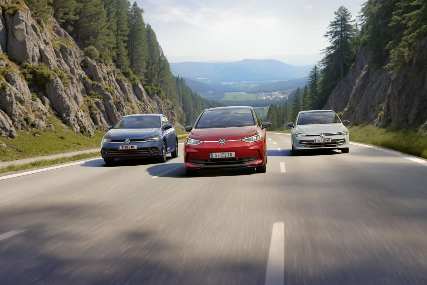 Drei VW fahren nebeneinander über eine dreispurige Landstraße. auf der mittleren Spur fährt ein roter VW ID.3, links daneben ein weißer Golf und rechts ein blauer Polo. Die Straße führt durch eine Waldlandschaft und ist umgeben von Felswänden und die Sonne scheint. 