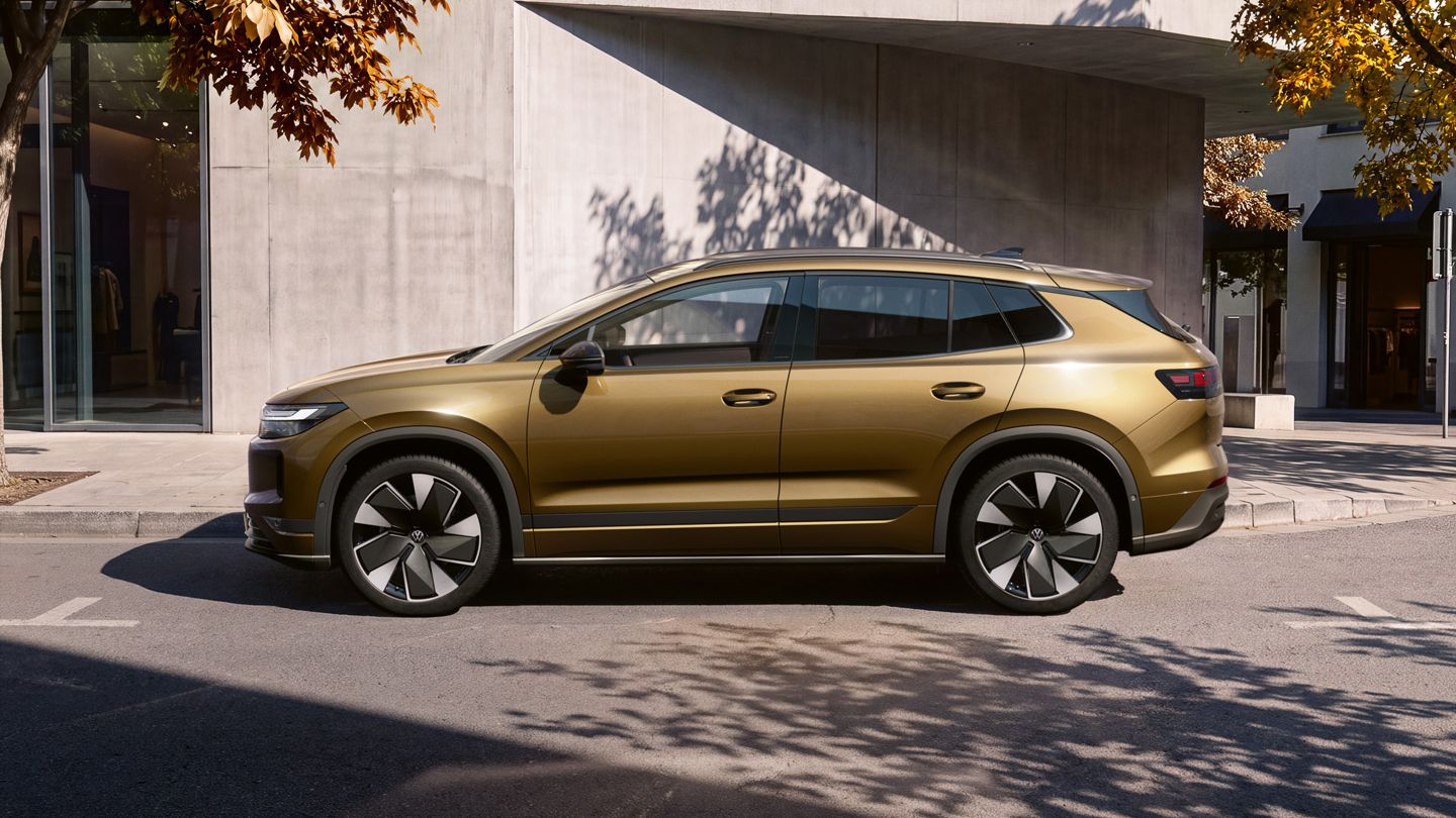Seitenansicht eines oliv-bronzefarbenen VW ID.4 Reskin geparkt vor einem modernen Wohngebäude.
