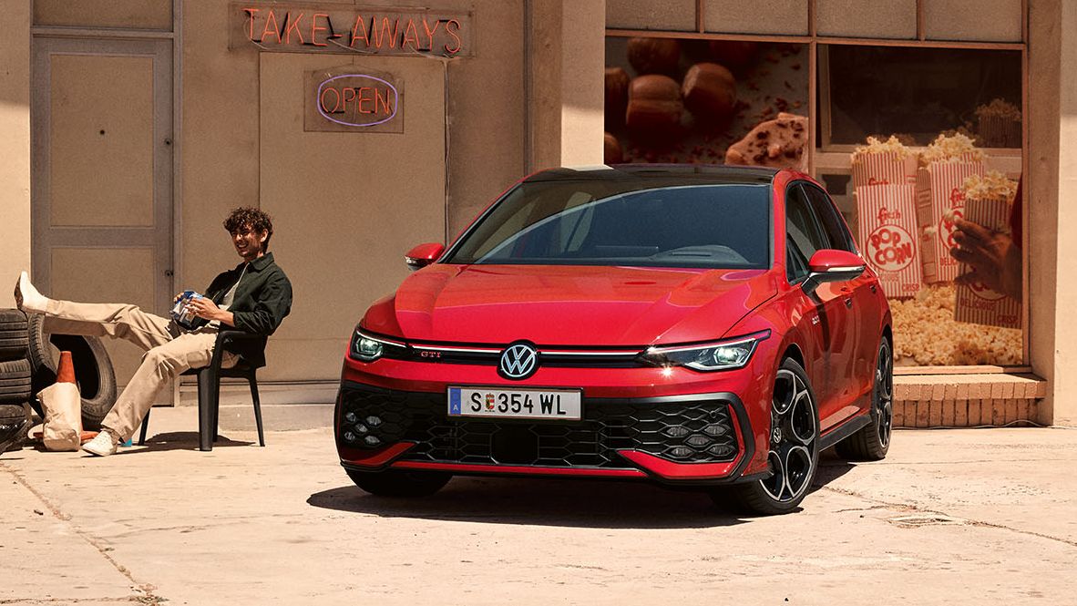 Zwei Personen sitzen auf Plastikstühlen neben einem Golf GTI, dahinter befinden sich ein Shop und ein Restaurant.