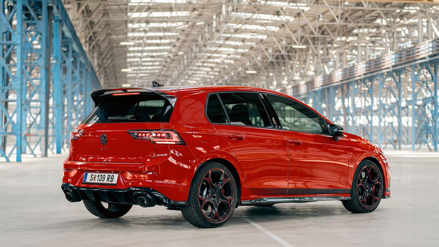 Ein roter VW Golf GTI steht in einer großen, lichtdurchfluteten Industriehalle. Das Auto ist von hinten und leicht seitlich zu sehen. Die schwarzen Räder und der Heckspoiler fallen deutlich ins Auge.