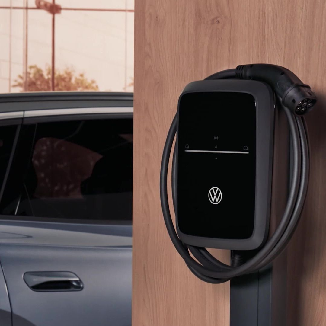 Ein Volkswagen ID..7 Tourer ist neben einer Ladestation geparkt. Die Ladestation besitzt ein schwarzes Bedienfeld mit einem VW-Logo und einem darum gewickelten Ladekabel.