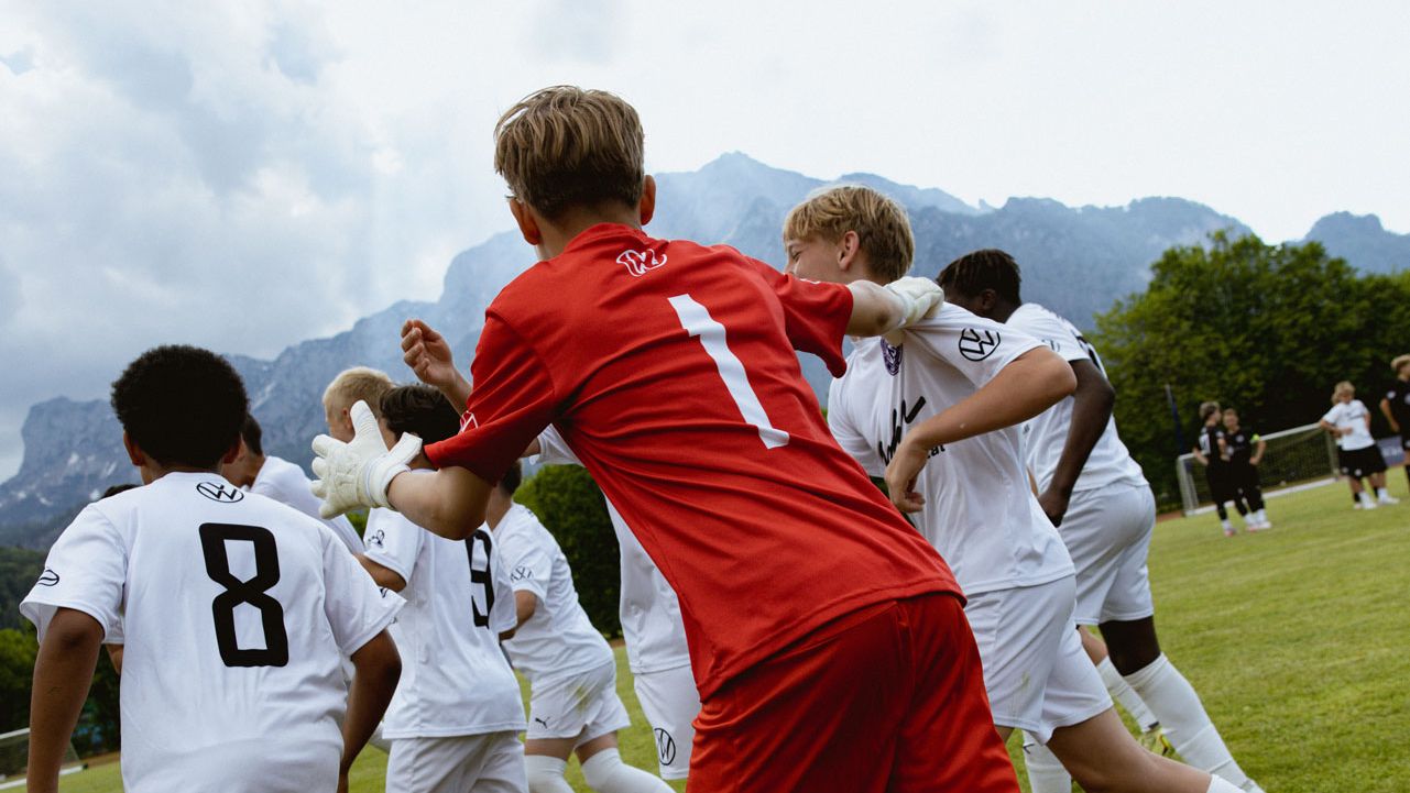 Mehrere Jugendliche in weißen Trikots jubeln auf einem Fußballfeld, während ein Spieler im roten Torwarttrikot mit der Nummer 1 zu ihnen läuft. Im Hintergrund sind Berge und ein bewölkter Himmel zu sehen. Die Szene wirkt dynamisch und feiert vermutlich ein erzieltes Tor oder einen Sieg.