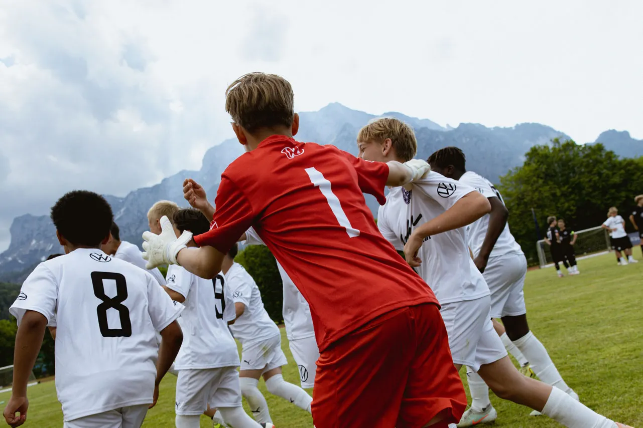 Mehrere Jugendliche in weißen Trikots jubeln auf einem Fußballfeld, während ein Spieler im roten Torwarttrikot mit der Nummer 1 zu ihnen läuft. Im Hintergrund sind Berge und ein bewölkter Himmel zu sehen. Die Szene wirkt dynamisch und feiert vermutlich ein erzieltes Tor oder einen Sieg.