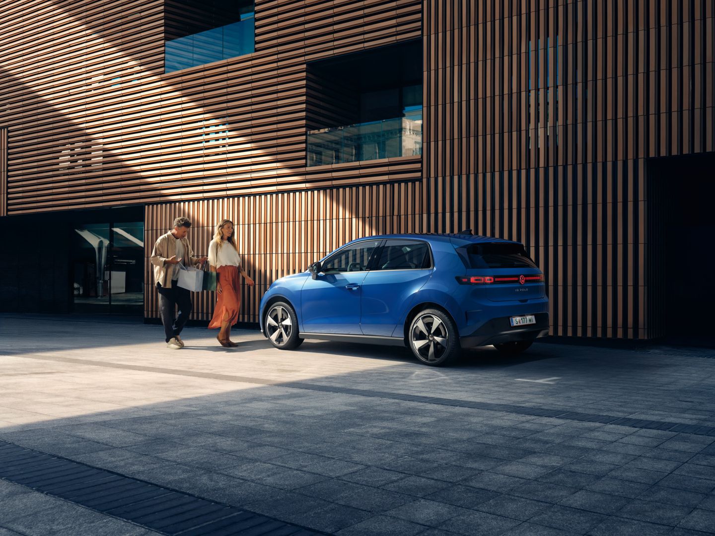 Ein blauer VW ID. Polo Style steht vor einem Gebäude mit Holzfassade. Ein Mann und eine Frau mit Einkaufstaschen gehen darauf zu