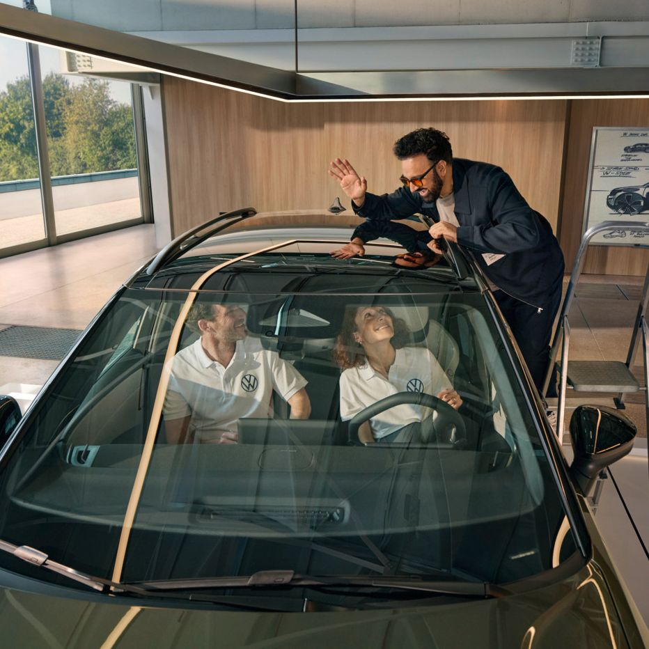 Ein Mann steht auf einer Leiter neben einem VW ID. Cross und winkt durch das Panoramadach den innen sitzenden Personen zu.