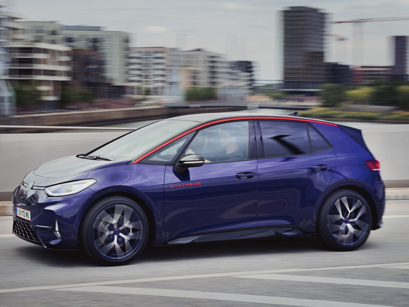 Ein blauer Kompaktwagen fährt auf einer Straße durch ein städtisches Gebiet. Die Gebäude im Hintergrund sind modern und mehrstöckig. Das Auto ist dynamisch in Bewegung und hebt sich durch seine markante rote Linie auf dem Dach hervor.