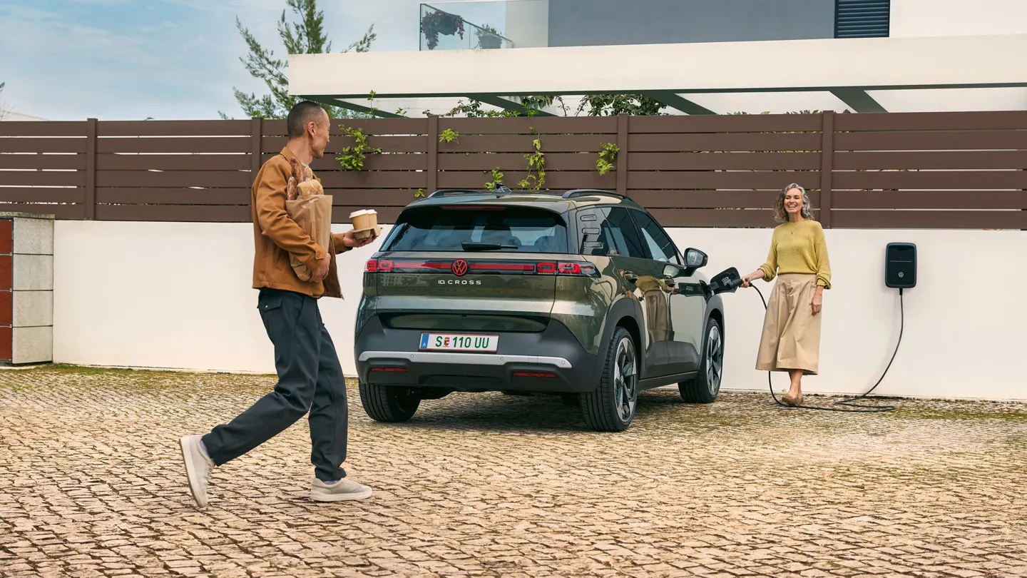 Eine Frau steckt ein Ladekabel einer Wallbox in einen grünen VW ID. Cross, ein Mann geht auf sie zu und hat Kaffeebecher in der Hand und Brot unter dem Arm.